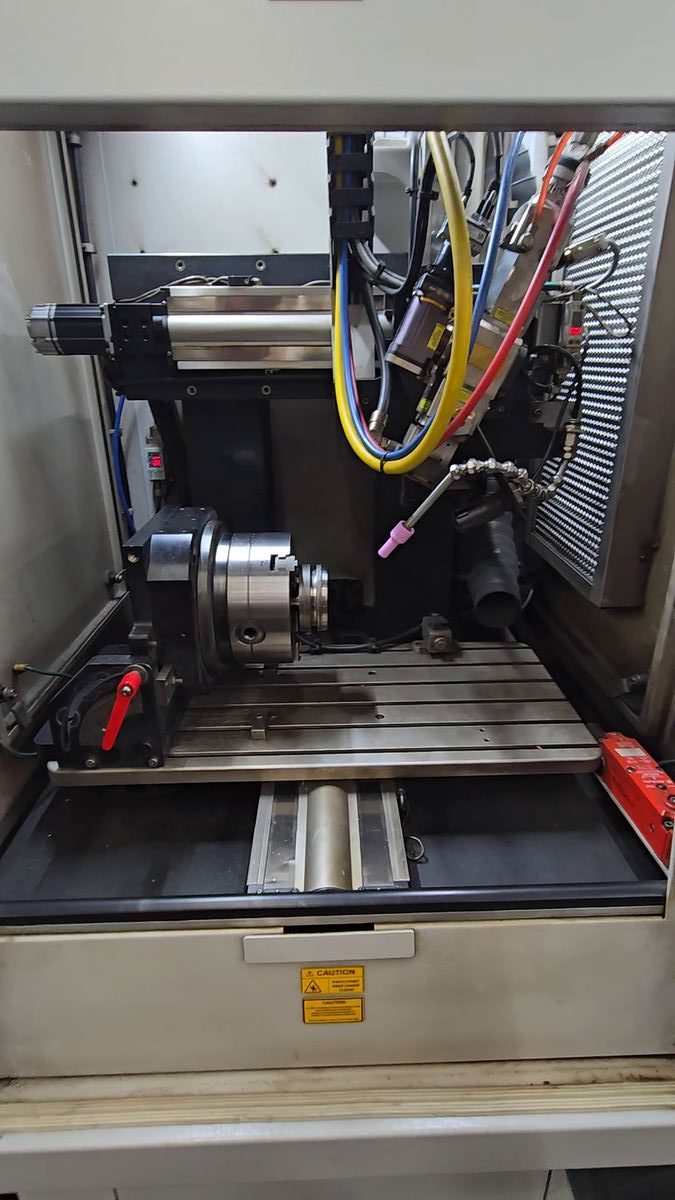 The work envelope with the enclosure door open: the chuck on its rotary indexer, the red tilt clamp lever, the pink ceramic nitrogen nozzle on its gooseneck, the service hose bundle and the T-slotted table.