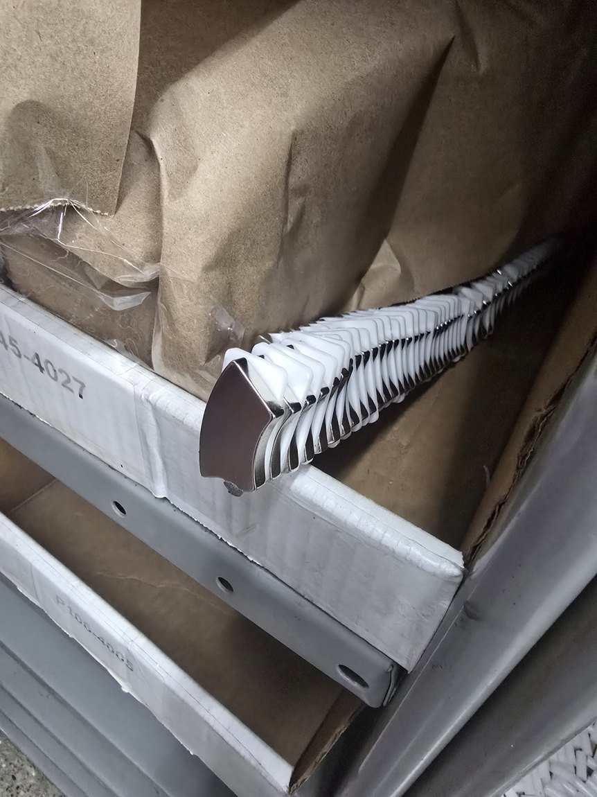A row of nickel-bright arc-shaped magnet segments interleaved with white plastic separators, in a kraft-lined shipping box.